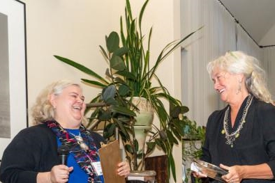 [ai] Two women laughing and exchanging awards in a warmly lit indoor setting with plants in the background.